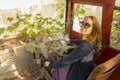 girl with blonde hair in sunglasses sits and smiles at the driver`s seat in an abandoned ivy-covered bus