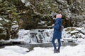 Girl in blue coat standing on a background of icy waterfall and rocks in a snow-covered coniferous forest. Winter cloudy day. Side Royalty Free Stock Photo