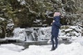 Girl in blue coat standing on a background of icy waterfall and rocks in a snow-covered coniferous forest. Winter cloudy day. Side Royalty Free Stock Photo