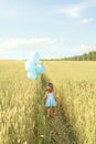 Girl in blue dress with balloons in hand runs among a large field of growing wheat on sunny summer day. Children`s holiday. Royalty Free Stock Photo