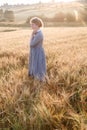 Girl in blue dress standing at dawn in field of ears in golden light of sun. hill and country landscape in background Royalty Free Stock Photo