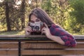 Girl in blue jeans and checkered shirt is sitting on the wooden bench and holding an old film camera in her hands Royalty Free Stock Photo