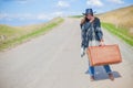 A girl in blue jeans, a poncho, a black leather hat with an old brown suitcase in her hands is on the road. Royalty Free Stock Photo