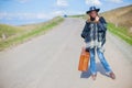 A girl in blue jeans, a poncho, a black leather hat with an old brown suitcase in her hands is on the road. Royalty Free Stock Photo