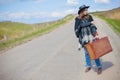 A girl in blue jeans, a poncho, a black leather hat with an old brown suitcase in her hands is on the road. Royalty Free Stock Photo