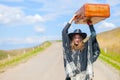 A girl in blue jeans, a poncho, a black leather hat with an old brown suitcase in her hands is on the road. Royalty Free Stock Photo