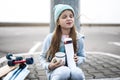 A girl in a blue sweater and hat was riding a longboard and sat down on the curb to drink hot tea from a thermos. Royalty Free Stock Photo