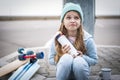 A girl in a blue sweater and hat was riding a longboard and sat down on the curb to drink hot tea from a thermos. Royalty Free Stock Photo