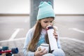 A girl in a blue sweater and hat was riding a longboard and sat down on the curb to drink hot tea from a thermos. Royalty Free Stock Photo