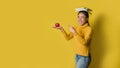 A girl with a book on her head and a red apple in her hand while the other hand points to the apple on yellow background. The Royalty Free Stock Photo