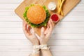 A girl with bound hands measuring tape reaches for a delicious burger. Light wooden background, healthy eating, top view