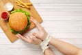 Girl with bound hands measuring tape reaches for a delicious cheeseburger. Light wooden background, healthy eating, top view