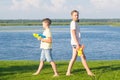 Girl and boy stand on green grass, their backs to each other and play with water pistols, against the blue sky and lake Royalty Free Stock Photo