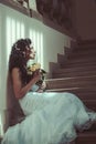 Girl with bridal makeup and hairstyle. Sensual woman with wedding bouquet. Woman with flowers sit on staircase. Bride in