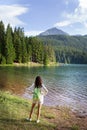 A girl with a bright backpack stands with her back near a mountain lake in the background. Positive young woman traveling on a Royalty Free Stock Photo