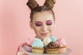 Girl with bright professional makeup and two buns hairstyle holds tray with multi-colored cupcakes on pink background. Bright