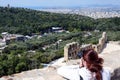 A girl with brown hair takes pictures of the Odeon. Ancient Greek ruins, ruins amidst lush green grass. Acropolis, Athens, Greece