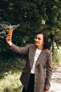 A girl in a brown jacket stay and takes a selfie. Portrait of a dark-haired girl in public park