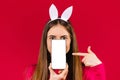 A girl with bunny ears holds a smartphone in front of her face and points to the screen with her index finger. Red Royalty Free Stock Photo