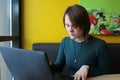 A girl with a calm expression sits, works at a laptop at a table in a cafe on a brown sofa against a yellow wall. He Royalty Free Stock Photo