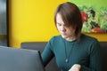 A girl with a calm expression sits, works at a laptop at a table in a cafe on a brown sofa against a yellow wall. He Royalty Free Stock Photo