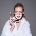 Girl on calm face wears bathrobe, grey background. Woman with face covered with foam holds straight razor in hand. Lady