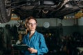 A girl car mechanic in the garage with a folder in her hands in goggles and describes the problem with the engine