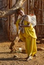 A girl carrying a jerry can of water on her back rests her head on a tree to rest.