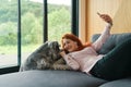 Girl with red hair and charming smile lying on sofa and making selfie with her pet