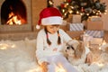 Girl child celebrates Christmas with Pekingese dog at home near Christmas tree, sitting on floor, hugging her pet and looking at Royalty Free Stock Photo