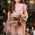 Girl child in a pink dress holding a Christmas gift in her hand, on the background of the New Year fir tree with decorative toys Royalty Free Stock Photo