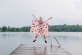Girl child in rain poncho with red maple leaves jumping on wooden lake dock. Canada Day holiday. Kid raising arms up under rain Royalty Free Stock Photo