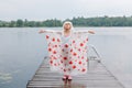 Girl child in rain poncho with red maple leaves standing on wooden lake dock. Kid rasing arms up under rain outdoors. Connection Royalty Free Stock Photo