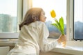 Girl child with yellow tulip flower, near the window sill looks out the window opening glass, sunny spring day