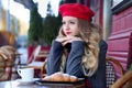 Girl in coat and a red beret with long curls sits thoughtfully near a cafe on a background of a red facade Royalty Free Stock Photo