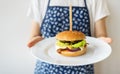 A girl cook in an apron shows serving a traditional hamburger with cheddar cheese and salad. Delicious and fast food.