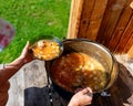 The girl cook outdoors puts soup with meat and vegetables borscht from a large cauldron pan on the wooden floor.
