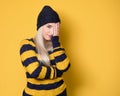 Girl covering face with hand. Shame, model wearing woolen cap and sweater, isolated on yellow background. Ashamed young woman