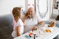 Girl with crown and wings applies powder onto father face in contemporary living room