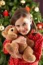 Girl Cuddling Bear In Front Of Christmas Tree