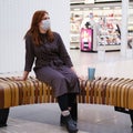 A girl in dark clothing and a medical mask sits on a bench in a shopping center