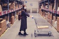 A girl in dark clothing and a medical mask stands with a cart in the warehouse