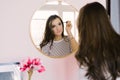 Girl with dark hair stands in front of a mirror and does makeup, holding a brush in her hand, creating an image, beauty and style