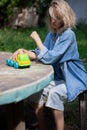A girl in a denim suit sits on a bench at a table outside in the garden and rolls a small toy car.