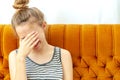 Girl in despair covers her face with hands sitting on a white sofa.