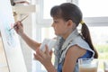 Girl diligently draws a picture on an easel in front of a large window