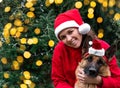 Girl with dog on background of bright yellow lights garlands. Young cute European woman in red Santa hat hugs black red German Royalty Free Stock Photo