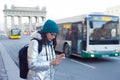 Girl in a down jacket and hat stands at a public transport stop and uses a smartphone. Wait for the bus.