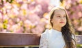 Girl on dreamy face sits on bench, sakura tree on background, defocused. Cute child with long beautiful hair enjoy sunny