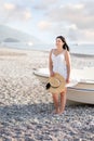 A girl in a dress and a hat in her hands stands by the boat on the seashore. Royalty Free Stock Photo